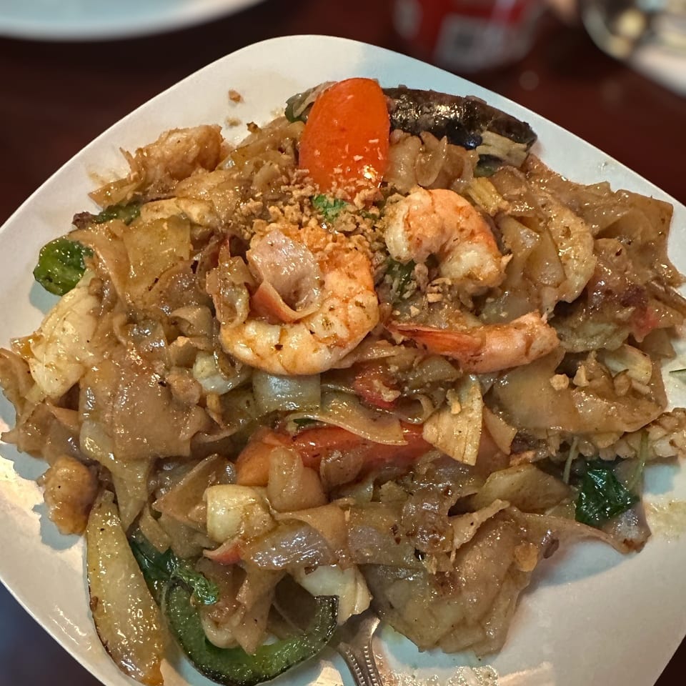 Plate of stir-fried noodles with shrimp, vegetables, crispy garlic, and chili peppers on a restaurant table.