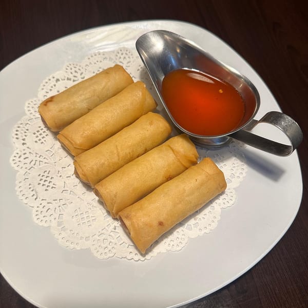 Six golden fried spring rolls arranged on a doily-lined plate with a metal dish of sweet chili sauce.