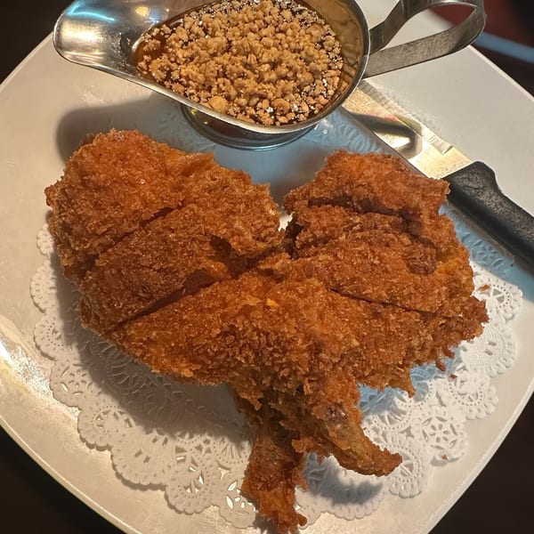 Golden-brown breaded fried pieces served on a decorative plate with a metal dish of dark dipping sauce and crumbs.